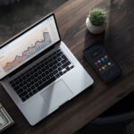 Mesa de trabalho com notebook prateado exibindo gráficos de analytics, notas de dólar empilhadas, smartphone e xícara de café dourada em ambiente de escritório minimalista
