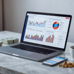 Notebook moderno exibindo gráficos de marketing digital ao lado de pilha de dólares e smartphone sobre mesa de mármore branca