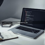 Notebook aberto sobre mesa branca exibindo código HTML e CSS na tela, ao lado de caderno com rascunhos de wireframe de site e xícara de café