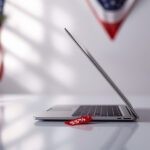 Computador portátil prateado moderno aberto sobre superfície branca com etiqueta vermelha de desconto e bandeira americana ao fundo desfocada
